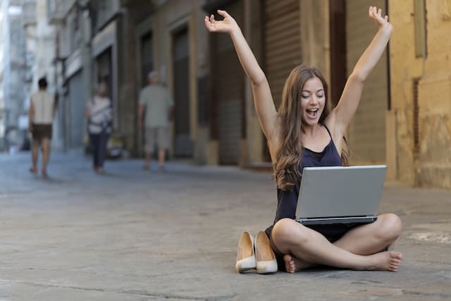 An independent consultant woman who is a digital nomad sits cross-legged on a street, hands in the air and a smile on her face as if cheering at her freedom to work anywhere she wants. Her laptop is open on her lap. Her shoes are next to her.