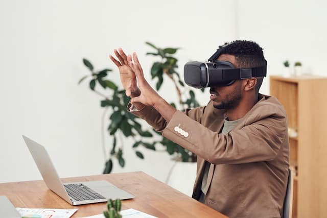 An independent consultant man at his desk in front of his laptop, wearing a virtual reality headset, with his hands in the air exploring an object in the metaverse.