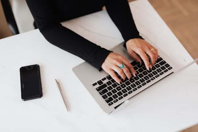 An independent consultant woman is at a computer keyboard crafting a proposal. Only her arms with her hands on the keyboard are shown. The keyboard is on a desk with a cellphone and a pencil.
