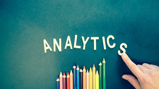 The index finger of a hand points to the word analytics which represent data and analytics reporting used by independent consultants.. Beneath the word analytics are multiple colored pencils of various lengths, oriented vertically and reminiscent of a histogram graph.