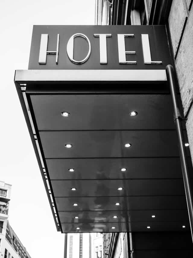 A picture of a hotel sign representing accommodation which independent consultants use when they travel. The sign is on an outdoor overhang projecting from a hotel which says "HOTEL." Looking up there are bright circular lights under the overhang.