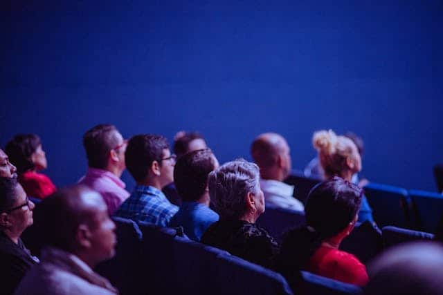 Independent consulting attending a professional conference meeting to which they have traveled. They are seated in theater-style seats in a darkened room, all looking slightly upward.