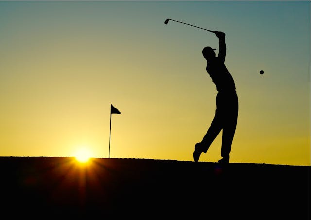 Consultant using deliberate practice to improve his golf game. He is standing with his body arched at the end of a golf swing. He is silhouetted against the sunset and the flag of the green is behind him. The ball is in the air.